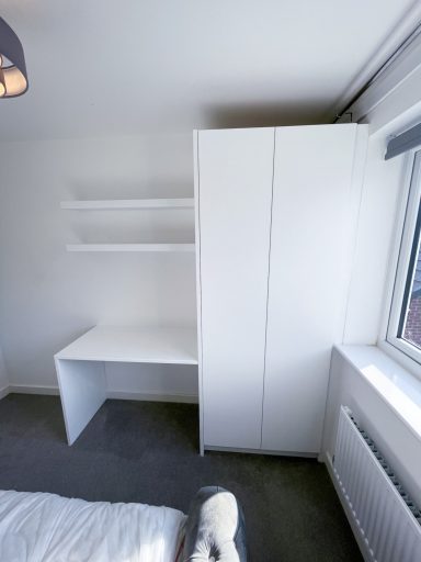 A minimalist bedroom featuring a white wardrobe, desk, and shelves by a window.